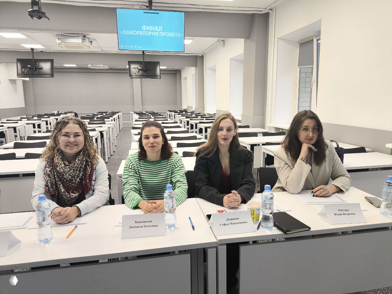 Фотография жюри за столом: четыре эксперта сидят в ряд, перед ними бейджи и бутылки воды в аудитории ТГУ.