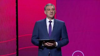 Мужчина в деловом костюме с планшетом на сцене на фоне ярко-розового экрана — официальное выступление в деловом контексте.