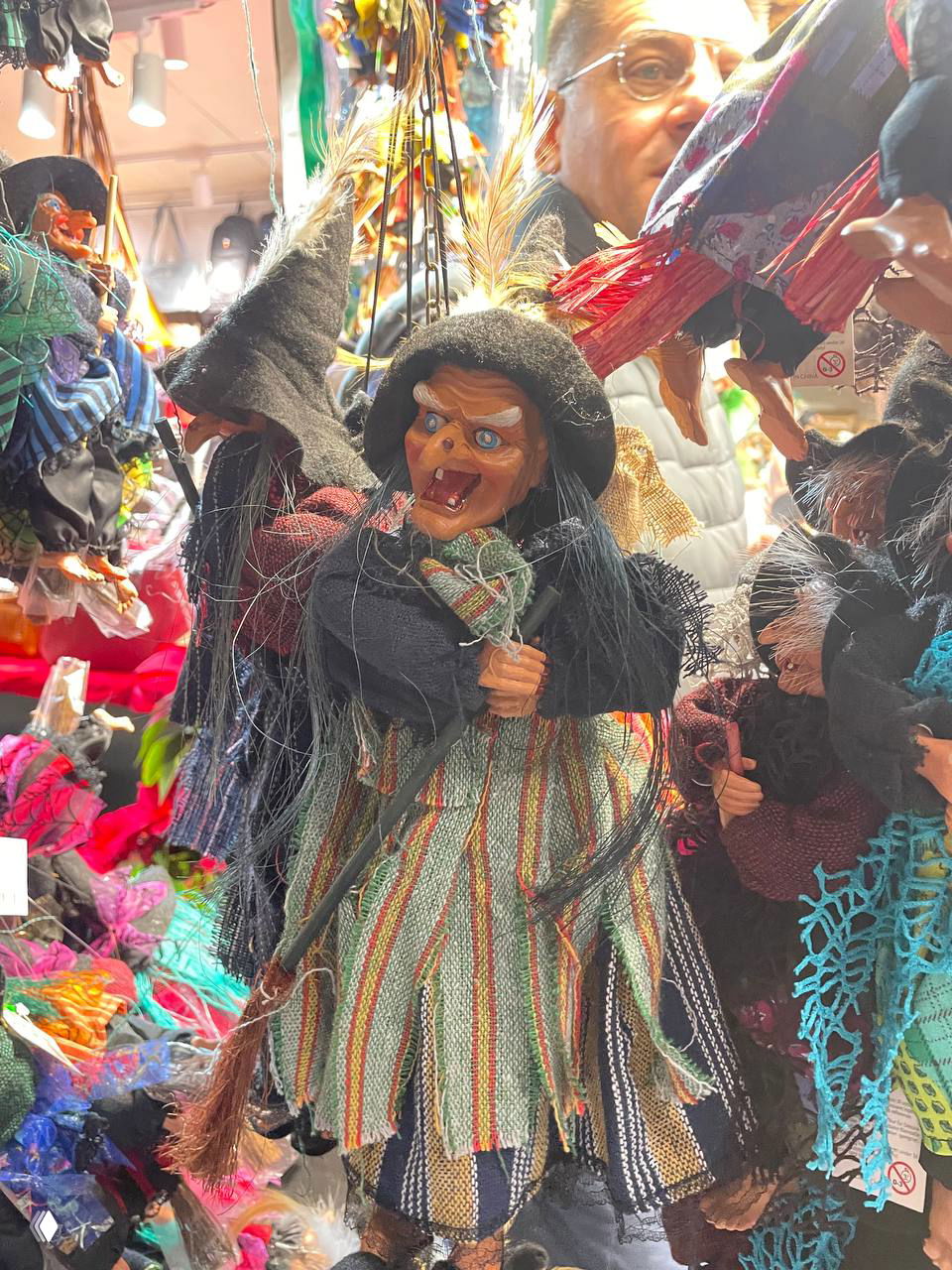 Крупный план чёрной ведьмы-куклы среди других тряпичных кукол и ярких мелочей на прилавке рождественского рынка в Риме.