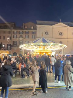 Карусель и плотная толпа людей на рождественской ярмарке в Риме, огни и праздничная атмосфера на площади вечером.