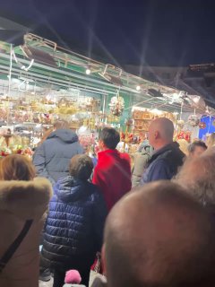 Ещё один кадр толпы на рождественском базаре в Риме: люди рассматривают товары в ряду под ярким искусственным освещением.