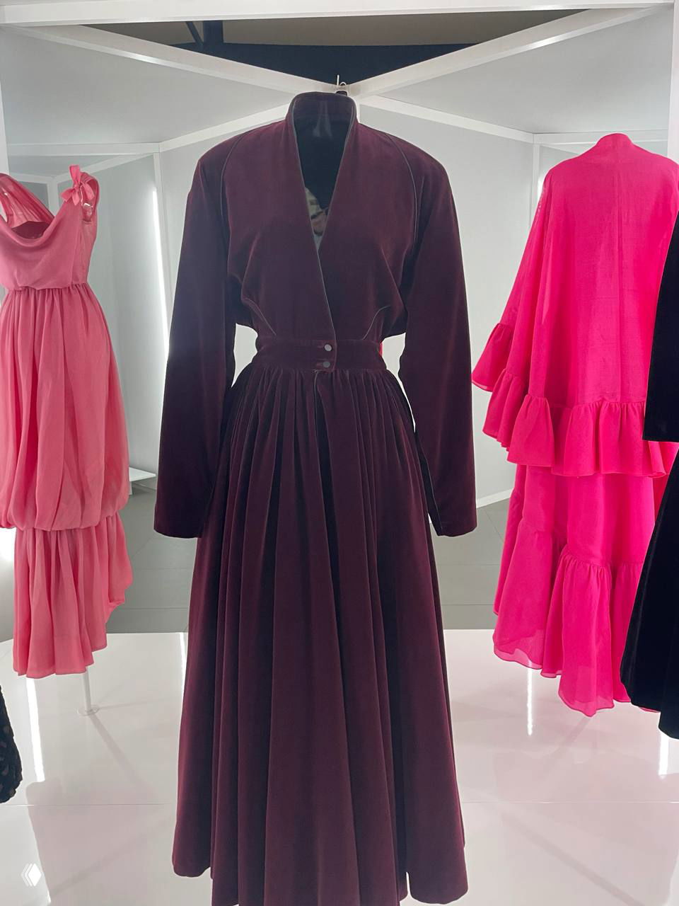 Бархатное платье тёмного винного цвета на манекене, рядом яркие розовые и коралловые наряды в экспозиционной комнате с зеркальной подложкой.