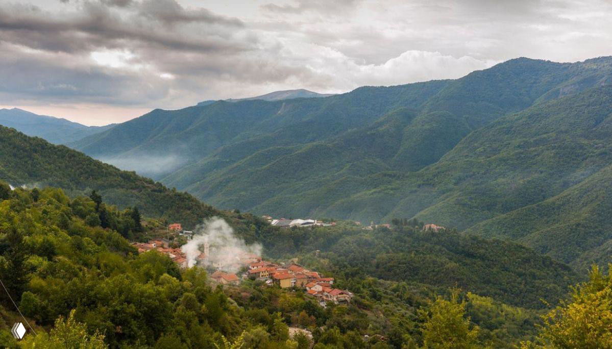 Горная панорама с небольшим селом в долине: зелёные склоны, дымок из труб и рассеянные постройки среди природы, типичная сельская сцена.