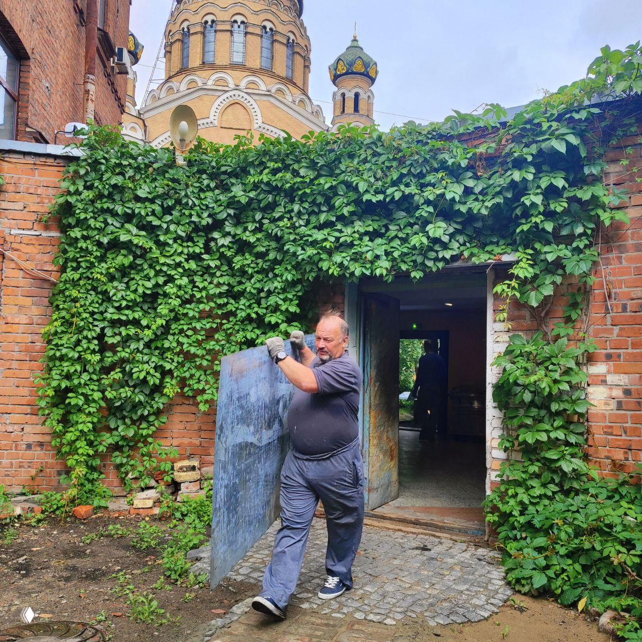 Мужчина-волонтёр несёт металлическую панель у кирпичного здания, на заднем плане виден купол православного храма; сцена из субботника и помощи общине.