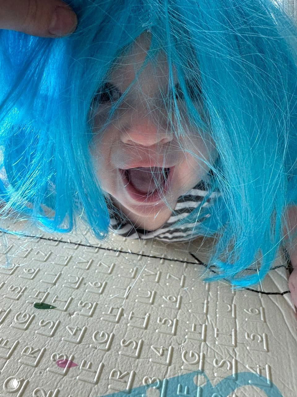 Малыш в ярком голубом парике с широко открытым ртом; крупный план, передающий искреннюю детскую радость и игру.