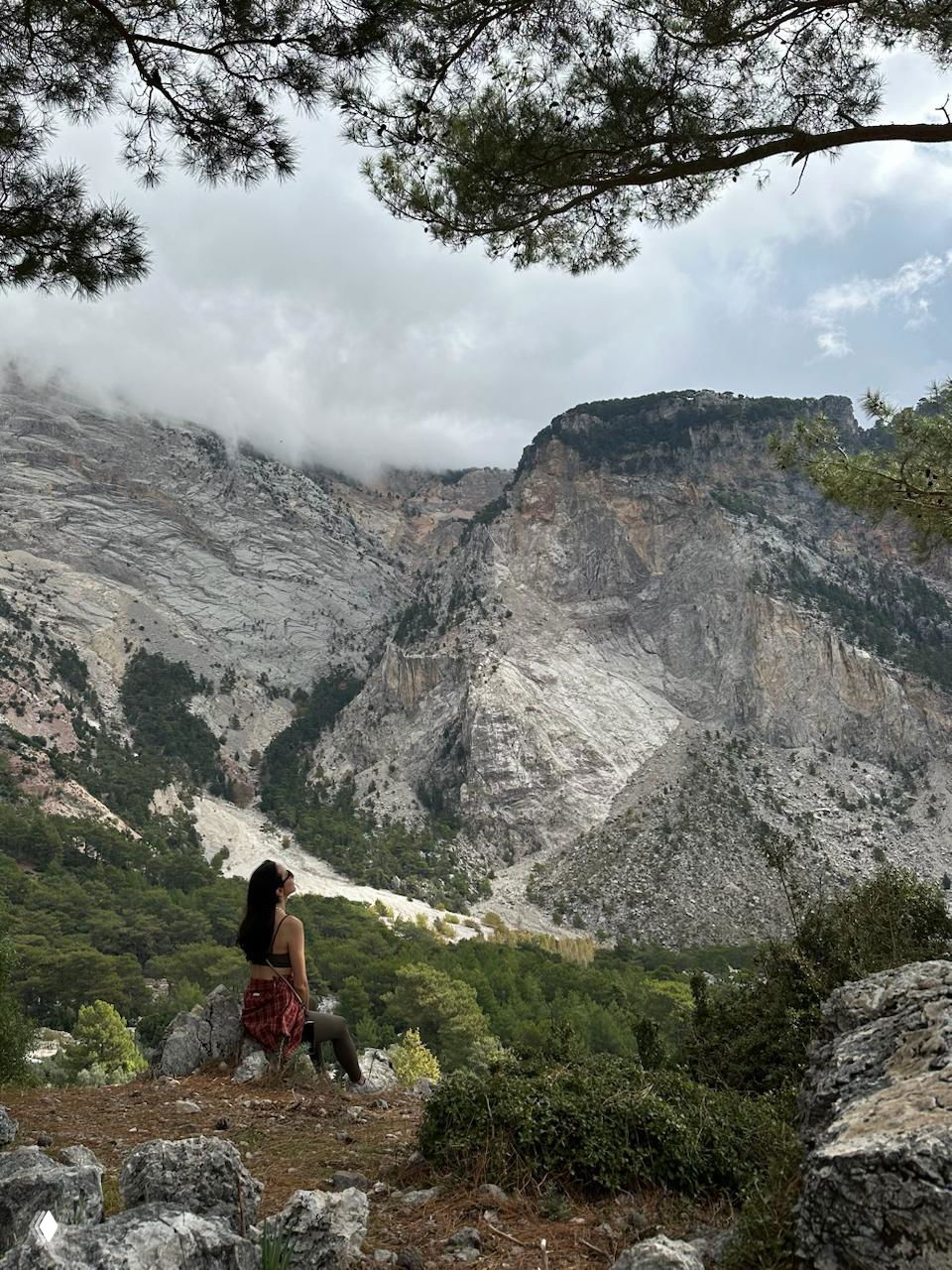 Человек сидит на камне у подножия скалы и любуется внушительными известняковыми стенами и низкими облаками над лесистыми склонами.