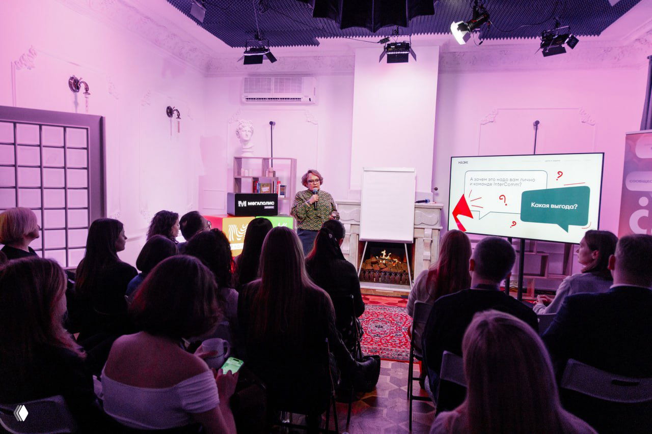 Зал с аудиторией на встрече НАЭКК: люди внимательно слушают спикера у экрана с презентацией, на заднем плане декор и камин