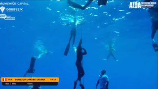 Подводный кадр с тренировкой по фридайвингу: ныряющий спортсмен у верёвки, рядом люди на поверхности, видна шкала глубин.