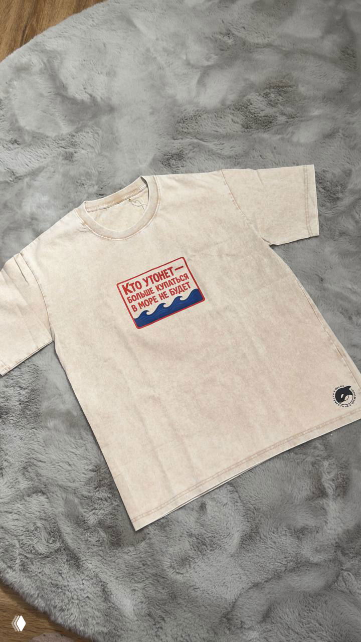 Светлая футболка с принтом «Кто утонет» красно‑синими буквами и волнами, разложена плоско на ковре — фото для анонса коллекции.