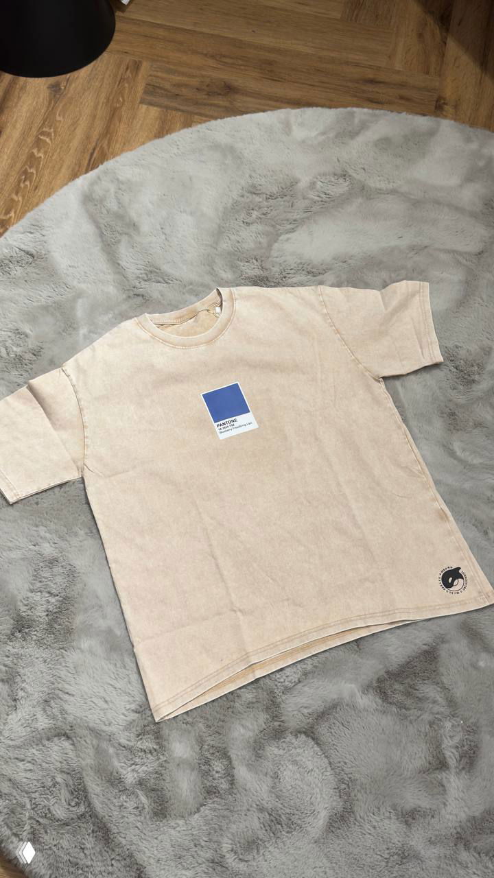 Бежевая футболка с квадратным принтом Pantone в центре, разложена на сером ковре — вид спереди для демонстрации дизайна.