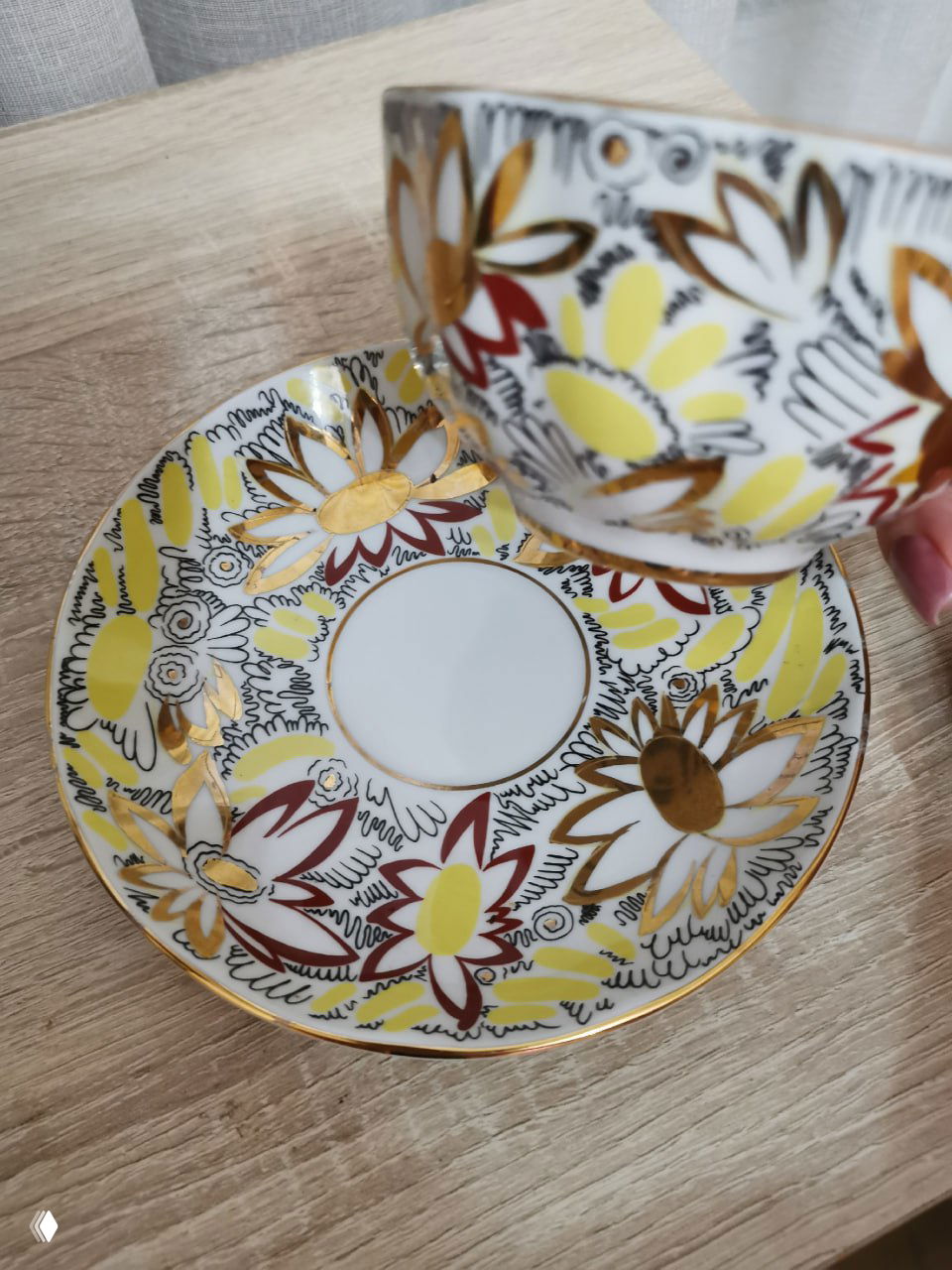 Крупный план фарфорового блюдца с жёлто-белыми цветами и золотым орнаментом, чашка приподнята рукой, видно текстуру столешницы и детали рисунка.