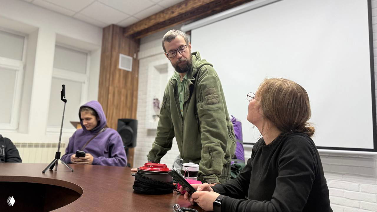 Мужчина в зелёной куртке общается с женщиной за столом на встрече; на заднем плане молодой человек в фиолетовом худи и штатив для съёмки — момент диалога с аудиторией.