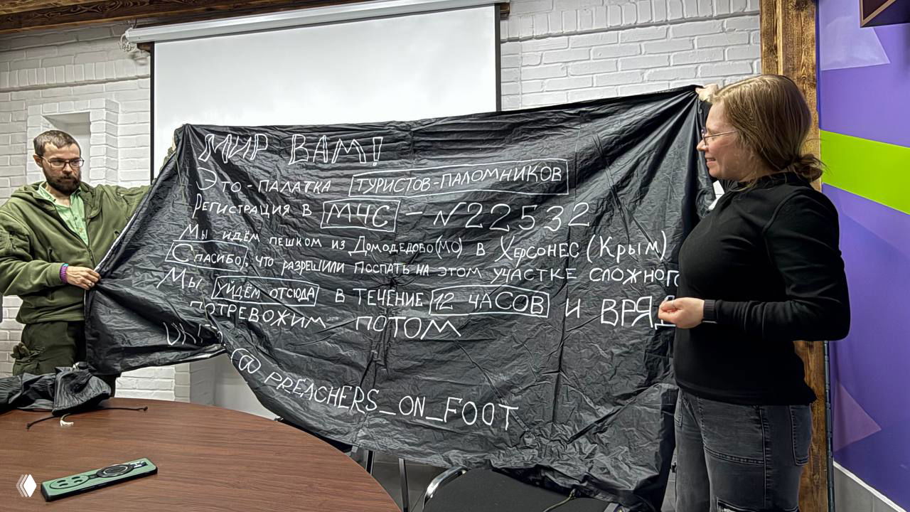 Двое участников демонстрируют чёрную ткань/палатку с белыми рукописными надписями о маршруте и регистрации в МЧС, видна подпись их канала preachers_on_foot.