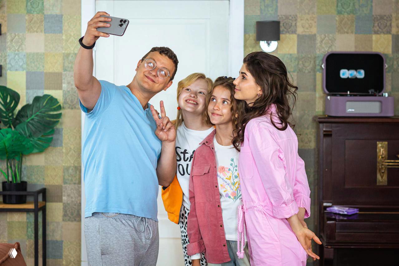 Группа персонажей делает селфи внутри комнаты: мужчина с телефоном держит кадр, рядом несколько девочек улыбаются — семейный и лёгкий момент из фильма.