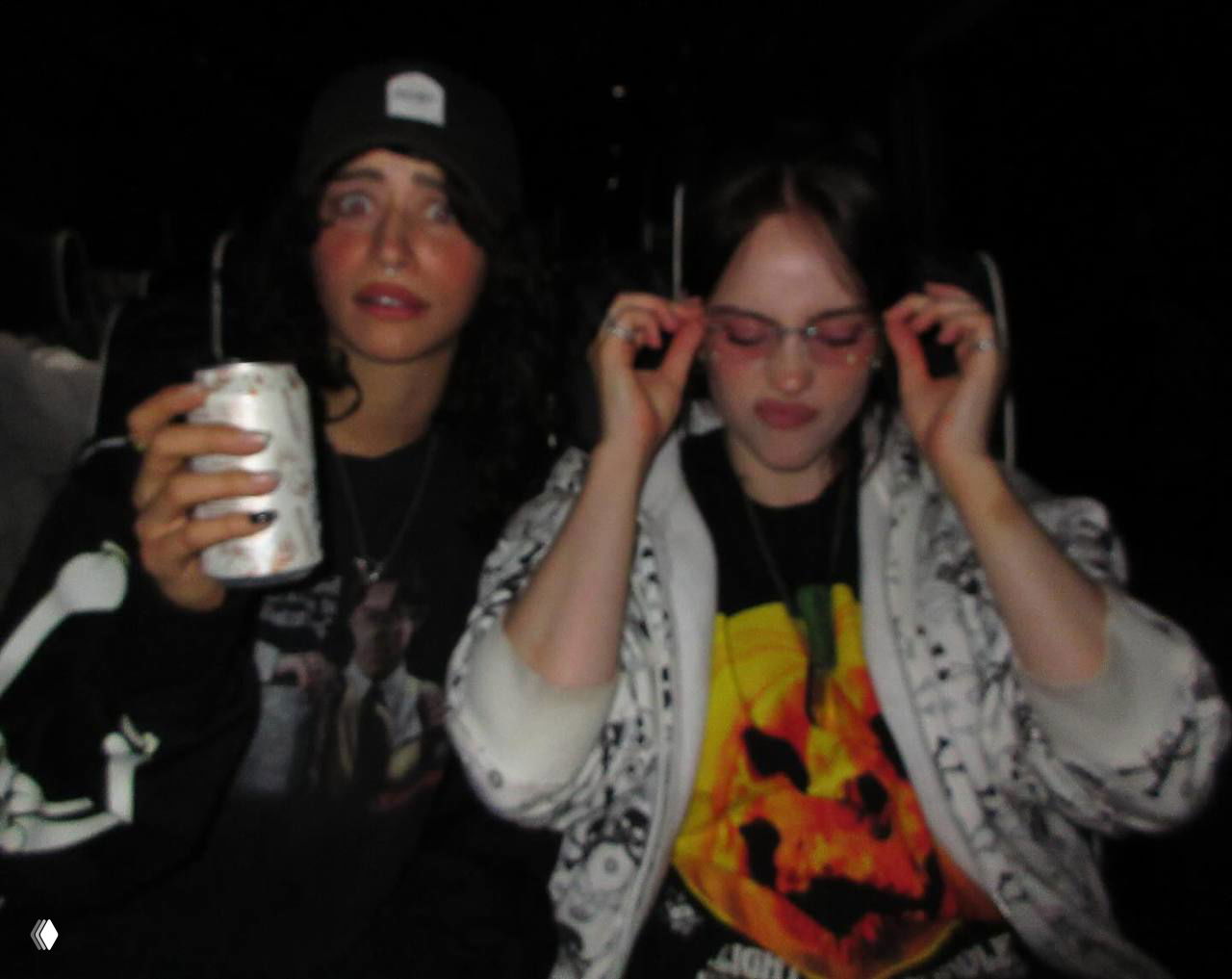 Ночная фотография: две молодые женщины в тёмной обстановке, одна держит напиток в банке, другая поправляет очки — атмосферный кадр вечерней тусовки.