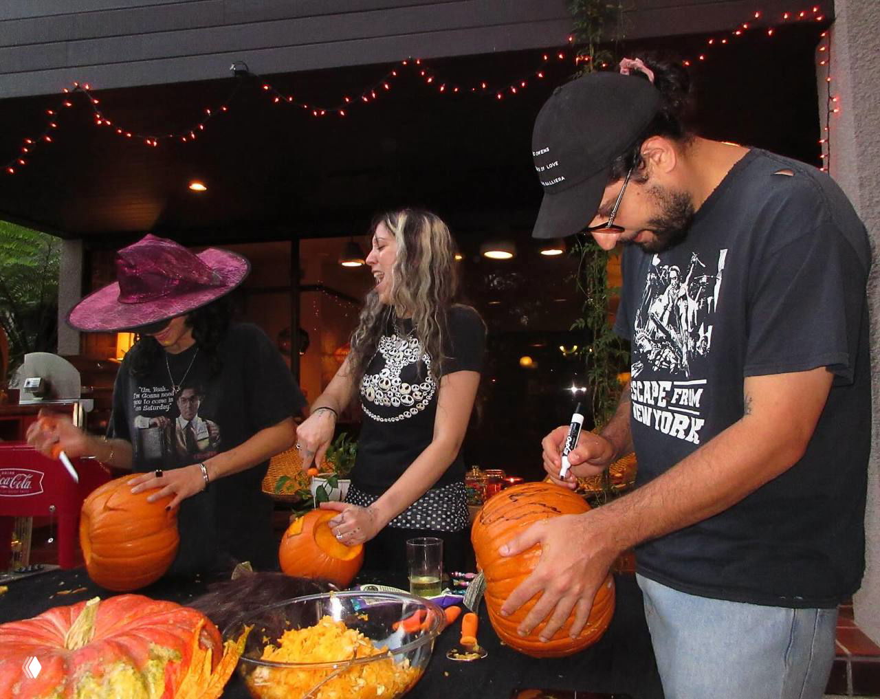 Группа людей за столом на открытой веранде вырезают тыквы; несколько резных и целых тыкв, инструменты для вырезания и весёлая подготовка к празднику.