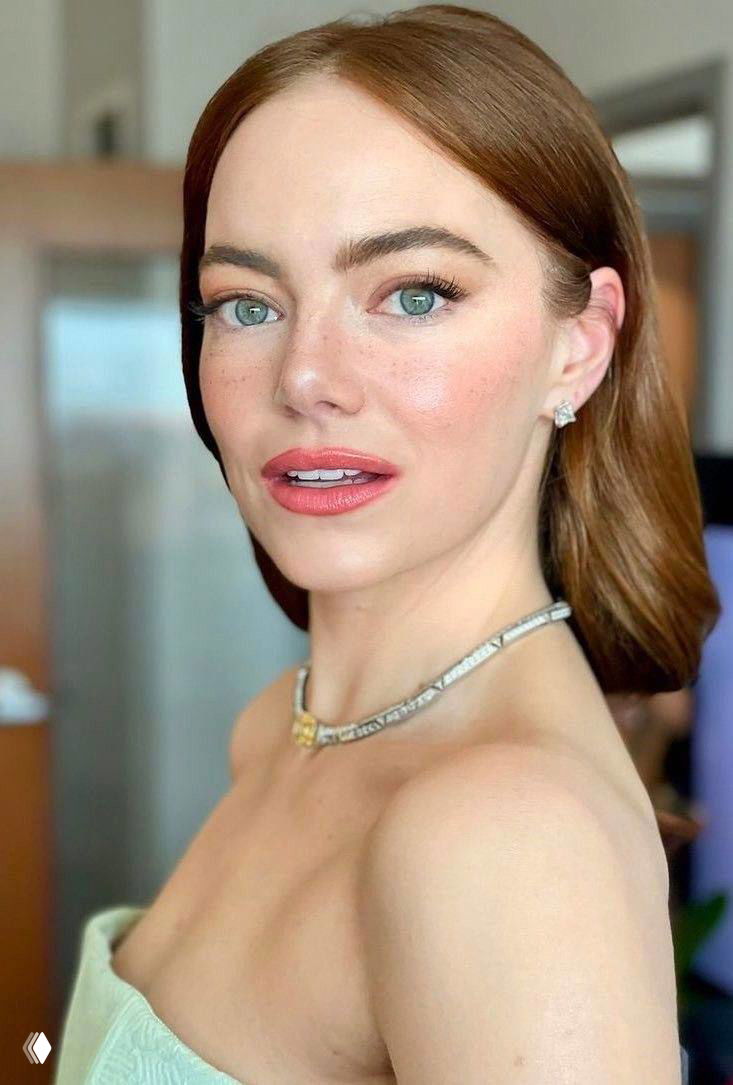 Крупный портрет Эммы Стоун с рыжевато-коричневыми волосами, светлым нюдовым макияжем и украшением на шее на размытом фоне интерьера.