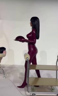 Бьянка Цензори в ярко-красном латексе в профиль, несёт поднос на сцене перформанса BIO POP; вокруг люди и объекты, стилизованные под мебель.