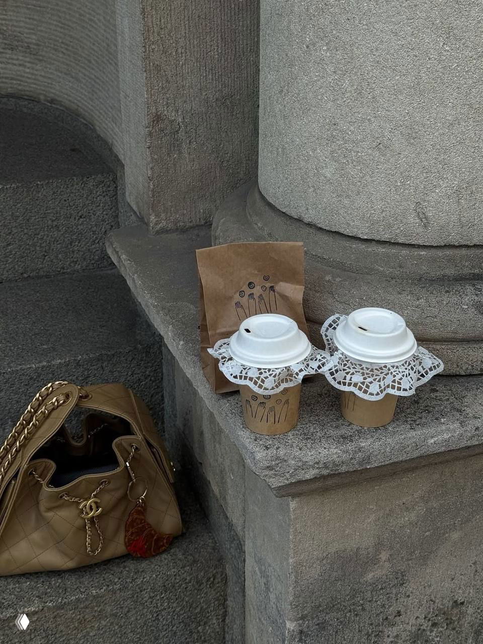 Два бумажных стаканчика кофе с кружевными подставками на каменной ступени рядом с сумкой — уличный аксессуарный кадр в стиле будуар.
