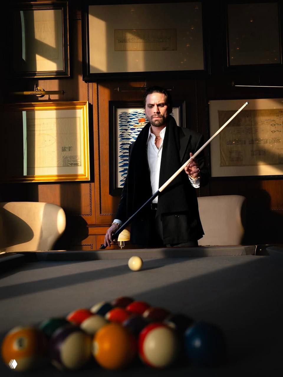 Мужчина в пальто с кием стоит у бильярдного стола в интерьере с картинами и тёплым солнечным светом, кадр в статичной позе.