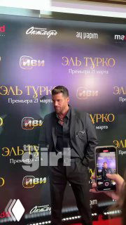 Джин Яман на красной ковровой дорожке в Москве у баннера премьеры: актёр позирует перед фотографами и смартфонами зрителей на фоне логотипов.