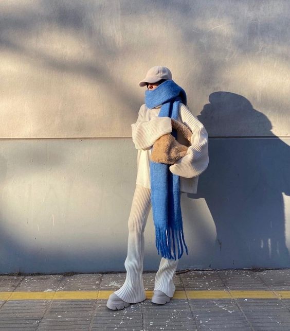 Модель в мягком голубом шарфе крупной вязки и светлой одежде на солнечном фоне — минималистичный зимний сет с акцентом на текстуру.