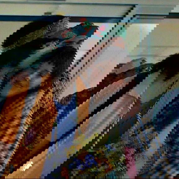 Съёмочный кадр: юноша целует девушку в щёку, они обнимаются на фоне людного пространства, в руках — букет цветов, тёплая сцена из эпизода.