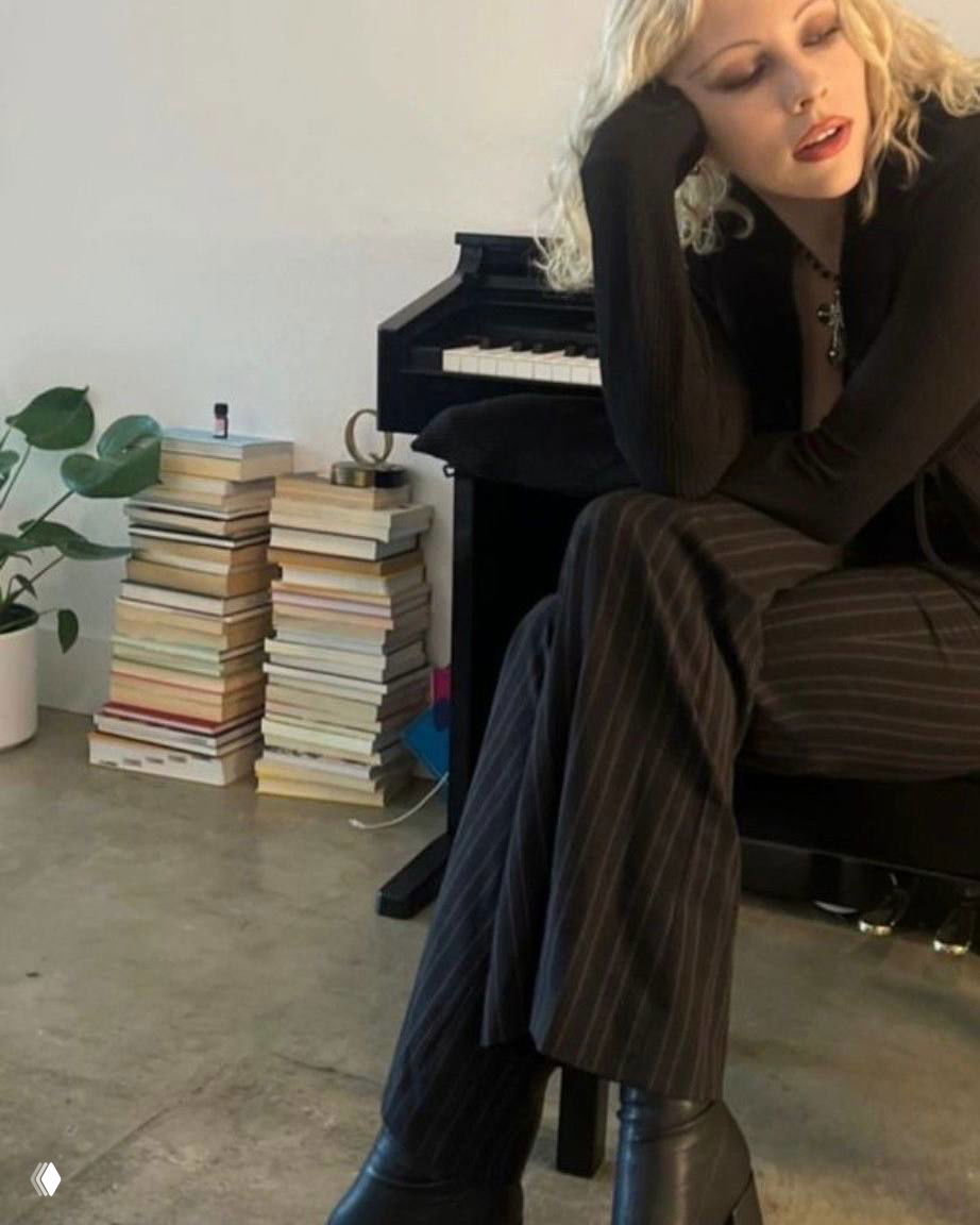 Женщина в тёмном комплекте сидит у пианино рядом со стопкой книг — интерьерный кадр с атмосфрой меланхоличного гламура.