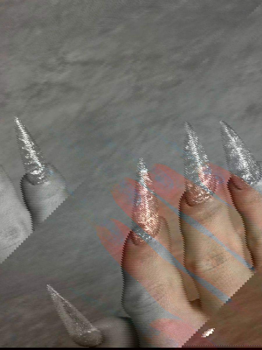 Крупный план очень длинных прозрачных стилетто‑ногтей с эффектом хрусталя и заострённой формой на светлом фоне.