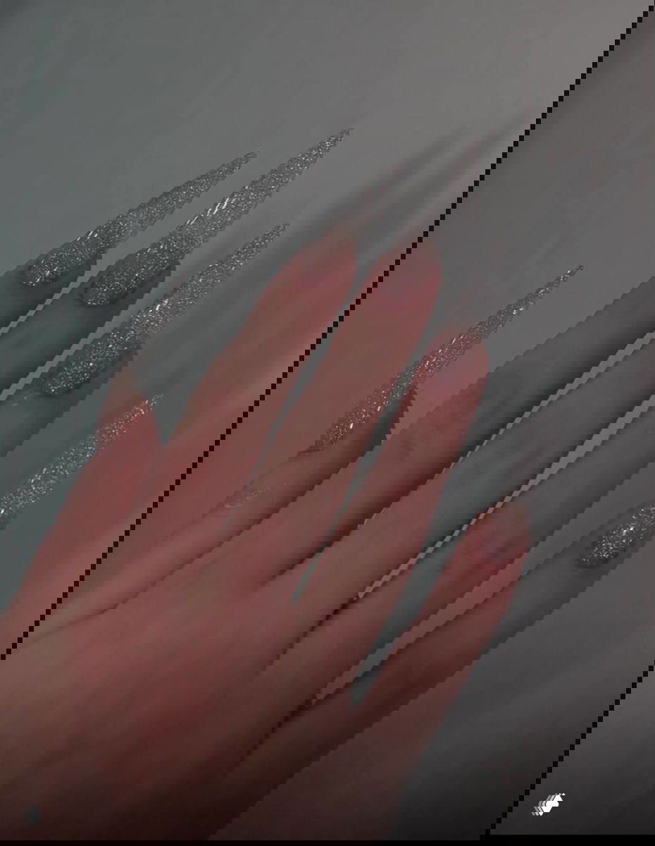 Рука с длинными прозрачными блестящими ногтями‑стилето, вид сверху на пальцы и мерцающую текстуру покрытия.
