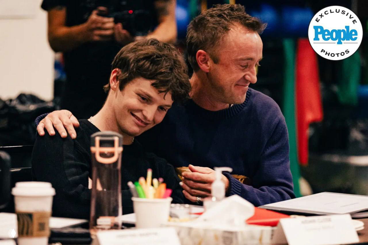 Том Фелтон на читке «Проклятого дитя» улыбается и обнимает коллегу; на столе чашки, реквизит и блокнот репетиции.