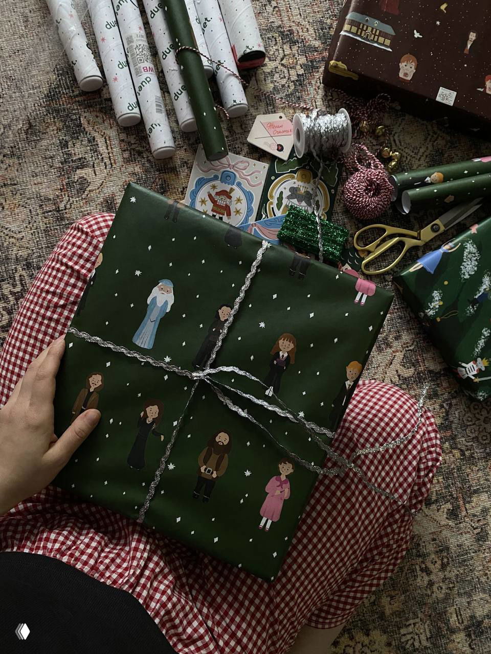 Рука держит подарок в тёмно‑зелёной упаковке с миниатюрными иллюстрациями персонажей на фоне клетчатых пижам и других материалов для упаковки.
