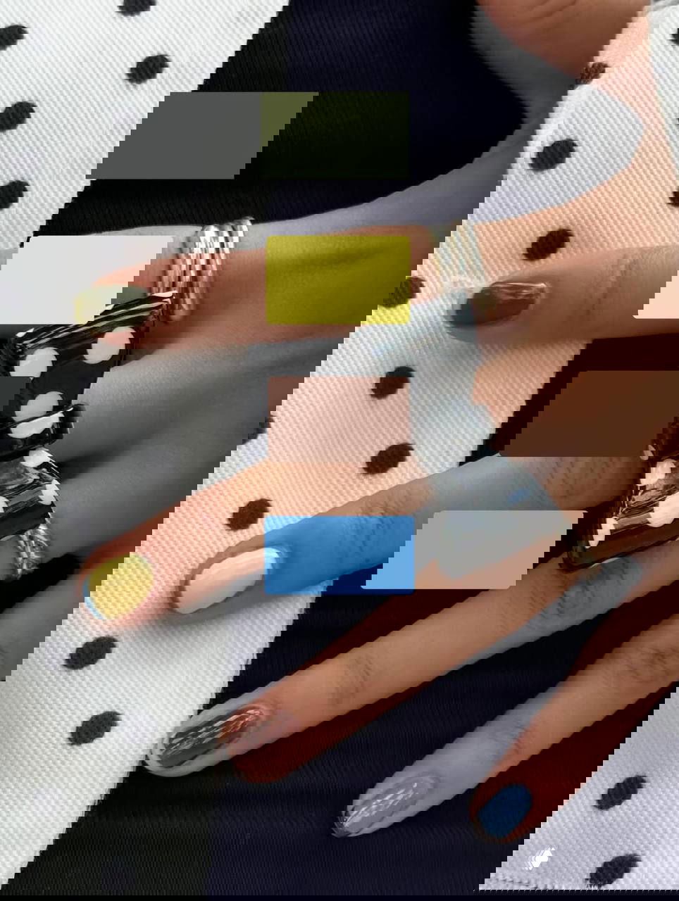 Крупный план рук с разноцветным маникюром: оливковые, жёлтые, коричневые и голубые акценты на коротких ногтях, заметное кольцо‑бантик и контрастный фон в горох.