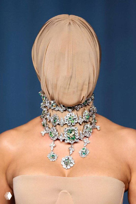 Фигура на красной дорожке с полностью закрытым бежевым чулком на голове и крупным драгоценным ожерельем с зелёными вставками.