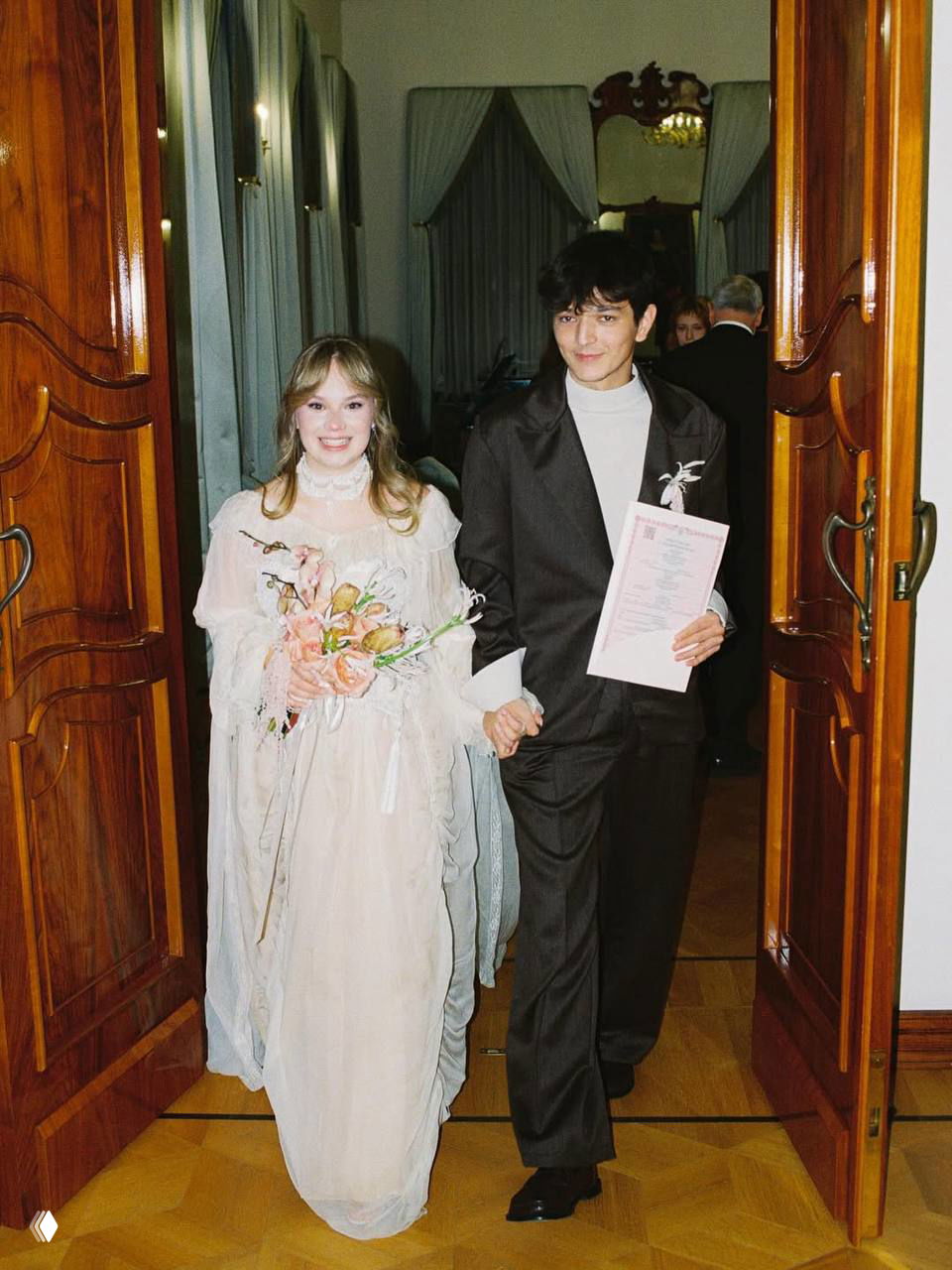 Невеста и жених выходят после регистрации: она в лёгком винтажном платье с букетом, он в тёмном костюме держит документ.