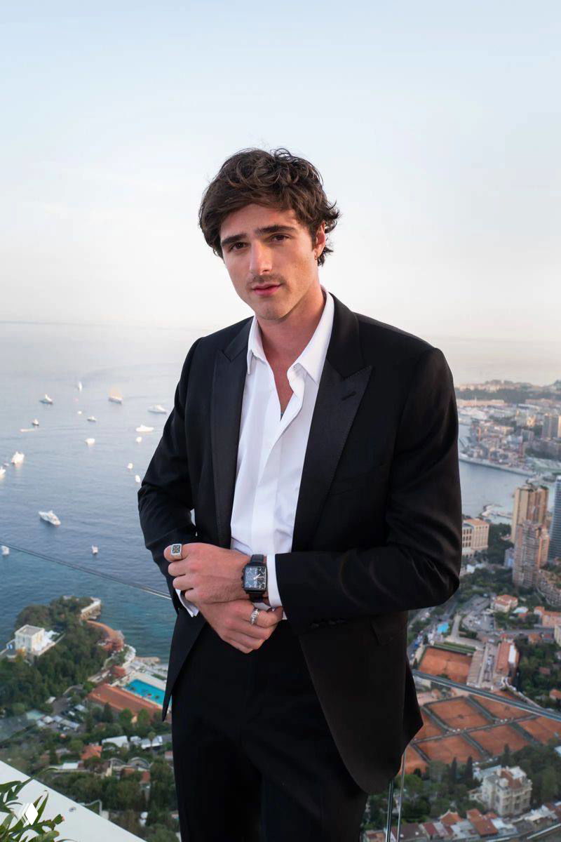 Портрет мужчины в чёрном смокинге на фоне прибрежного города с яхтами; вечерняя съёмка, элегантная поза и панорамный вид.