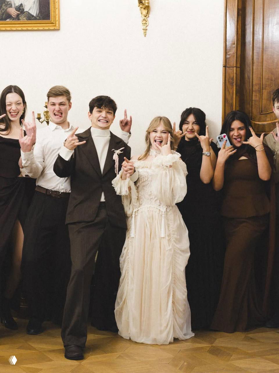 Групповое фото с гостями: жених и невеста в центре показывают жест «рок», вокруг друзья в вечерних нарядах, радостная и непринуждённая атмосфера.