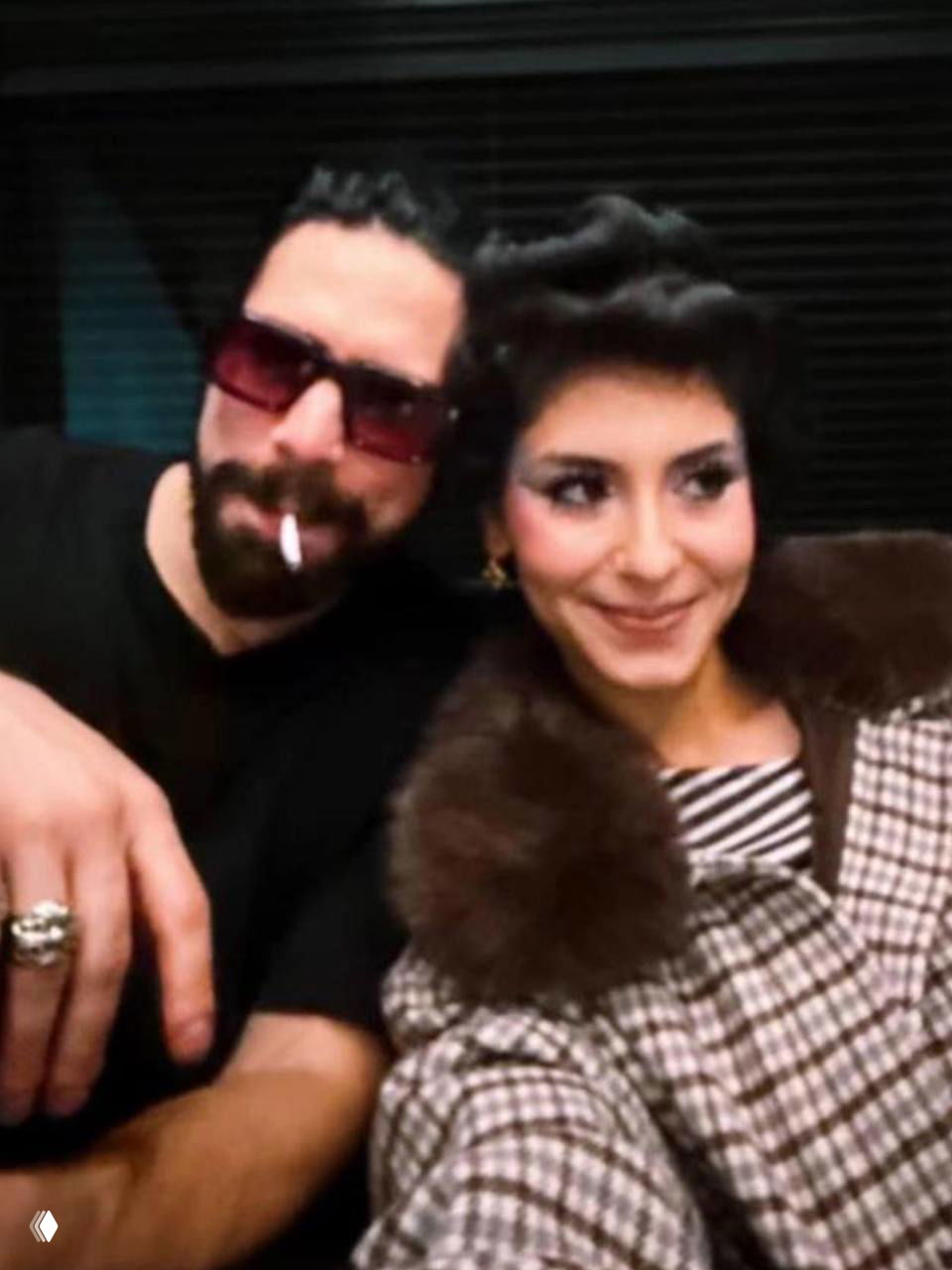 Два человека сидят рядом в тёплой обстановке: мужчина с бородой держит сигарету, женщина улыбается в ретро-образе, кадр в повседневной атмосфере.