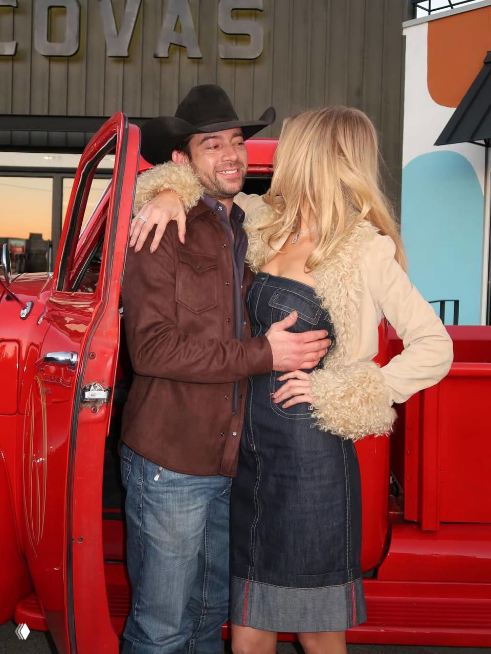 Пара обнимается у красного ретро-пикапа на улице: мужчина в ковбойской шляпе и женщина в джинсовом платье.