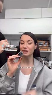 Вертикальный кадр: молодая женщина в серой толстовке держит предмет у рта на кухонном фоне; виден водяной знак TikTok, бытовая обстановка.