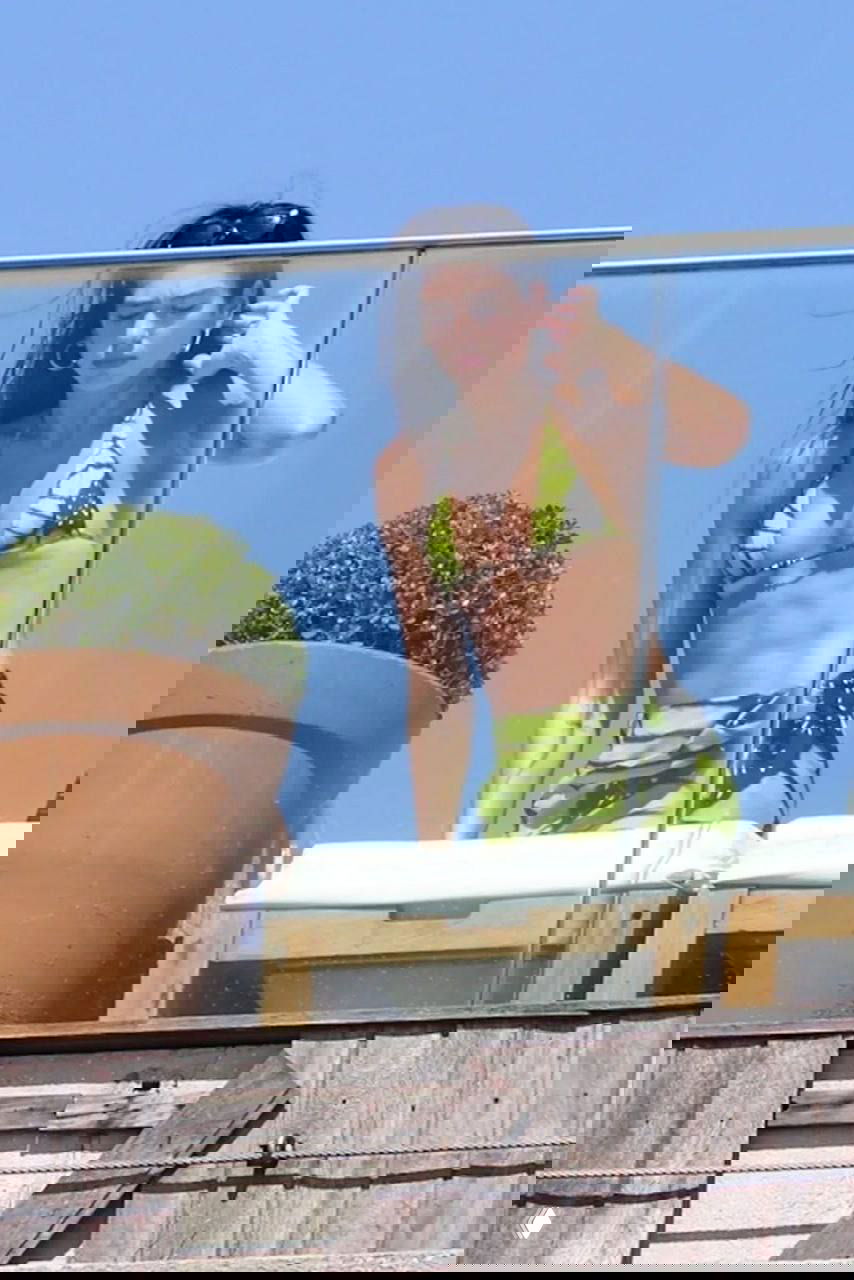 Женщина в зелёном купальнике с принтом крыла бабочки стоит за стеклянным ограждением на террасе у моря в ясный солнечный день.