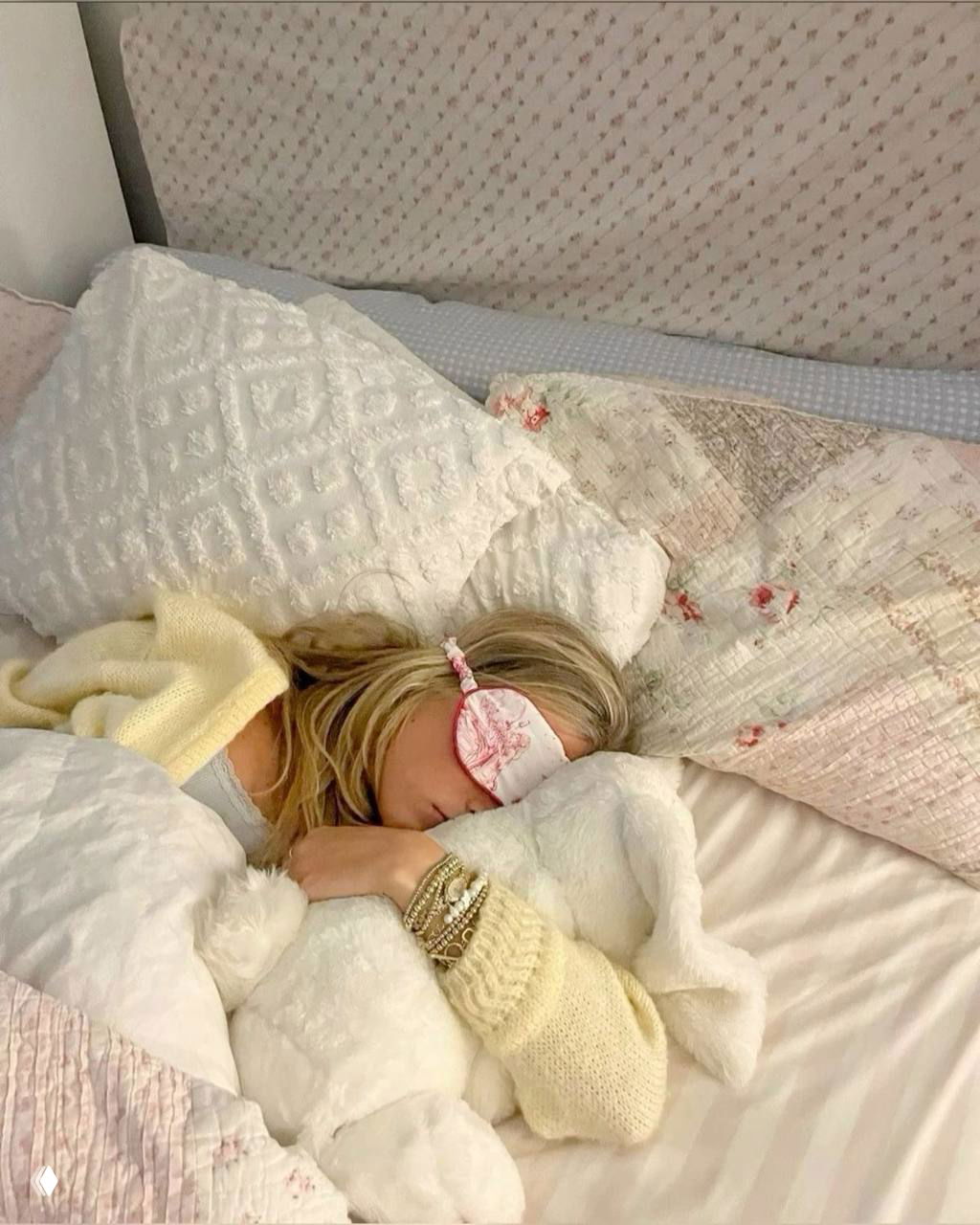 Девушка в маске для сна, завернутая в мягкие пледы и подушки на пастельной кровати — уютный кадр в предпраздничной атмосфере.