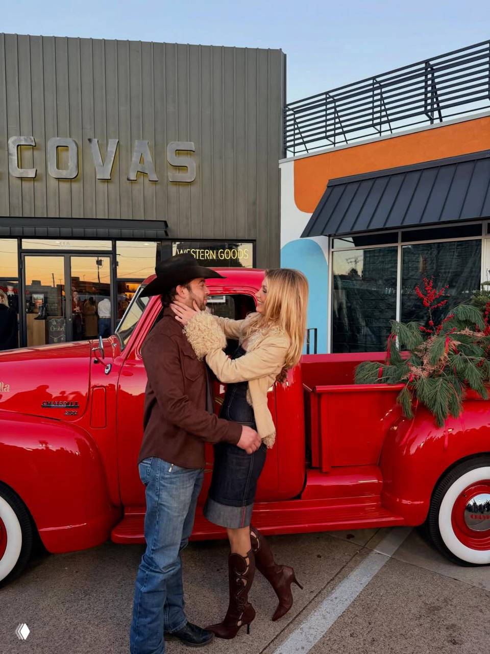 Пара целуется у красного фургона с праздничной зеленью; женщина в пальто с мехом и сапогах, мужчина в джинсах.