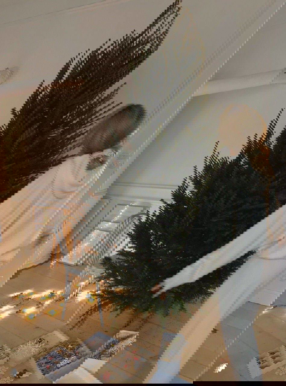 Женщина стоит у большой живой ёлки в светлой комнате; рядом коробки с игрушками и гирляндами — кадр подготовки к празднику.