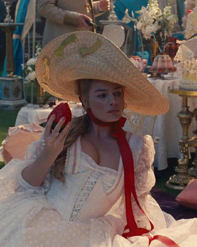 Актриса в широкополой шляпе держит в руке фрукт на праздничном приёме, видна роскошная сервировка и декор.