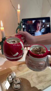 Две праздничные кружки с горячим шоколадом и маршмеллоу на деревянном столе; в фоне зажжённые свечи и экран с фильмом.