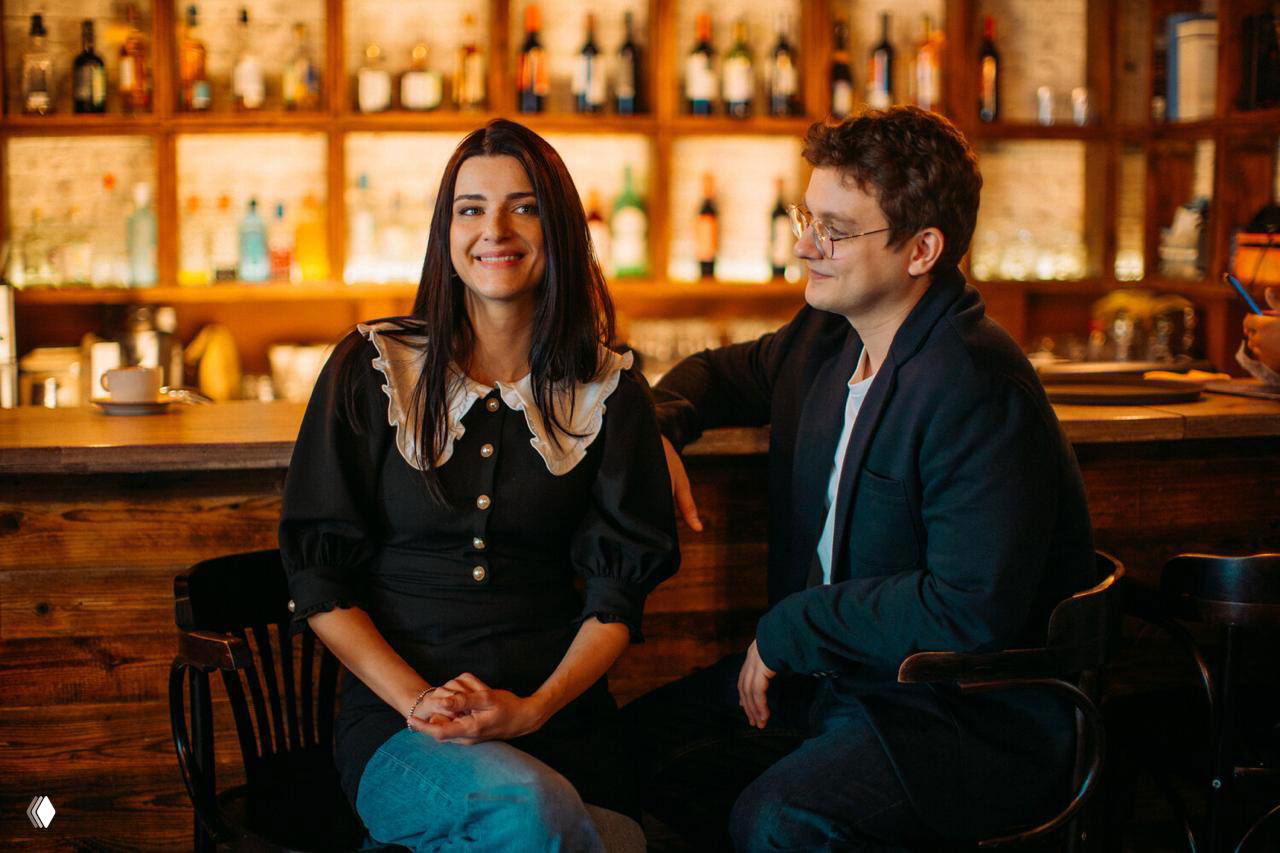 Кадр из фильма: молодая женщина в чёрном платье улыбается у барной стойки, рядом мужчина в очках наблюдает за ней, тёплая интерьерная сцена.