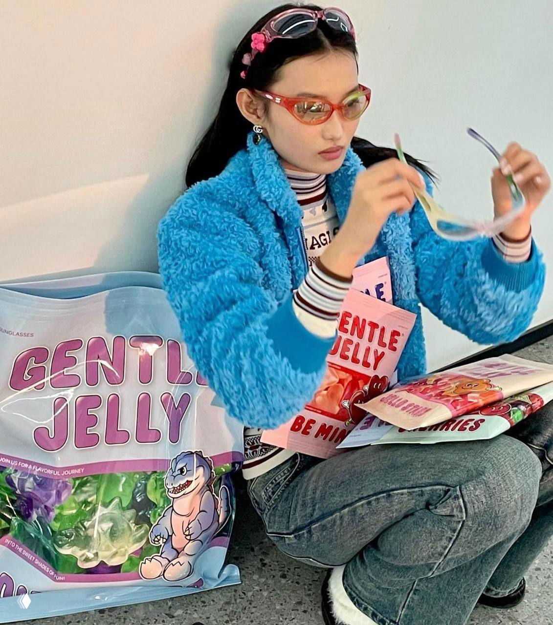 Девушка в голубой пушистой куртке примеряет разноцветные очки рядом с крупными упаковками «Gentle Jelly» и мармеладными аксессуарами — визуал трендовой эстетики.
