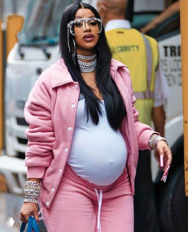 Карди Би в розовом спортивном костюме демонстрирует беременный живот на уличном кадре; подчёркнут её стиль и украшения.