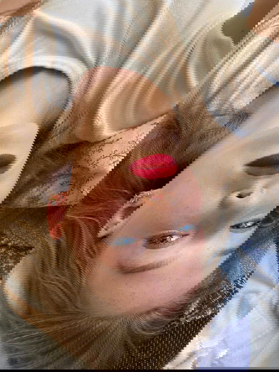 Девушка со светлыми/рыжеватыми волосами и яркой красной помадой, крупный план лица, видны естественные брови и текстура кожи.